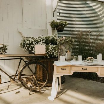 Rustic outdoor celebration setup with vintage bike, flowers, and elegant table setting under sunny skies.