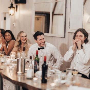 Wedding reception guests smiling and laughing around a rustic table with candles and wine bottles.