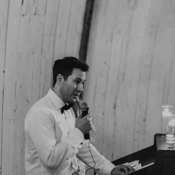 Man giving a speech at a wedding, wearing a white shirt and bow tie, speaking into a microphone.