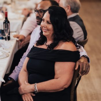 Smiling woman in black dress enjoying a wedding reception with friends at a beautifully set dinner table.