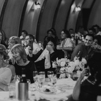 Guests seated at elegant wedding reception, smiling and emotional reactions, surrounded by decor and elegant table settings.