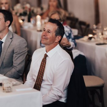 Man in white shirt and tie smiling while seated at a formal event, surrounded by other guests and tables.